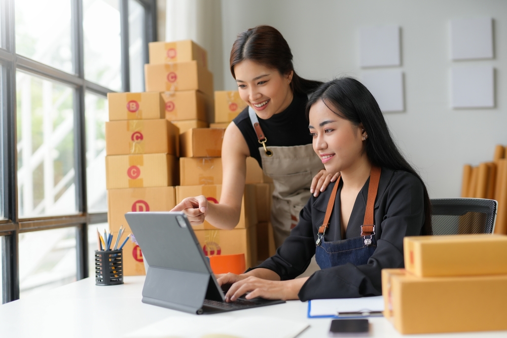 Two young asian women working together in a cozy home office, managing their online business surrounded by cardboard boxes and packages, using a tablet and computer for delivery logistics
